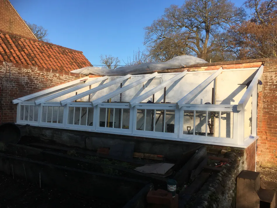 Long white wooden greenhouse with multiple glass panels attached to a brick wall under a clear blue sky.