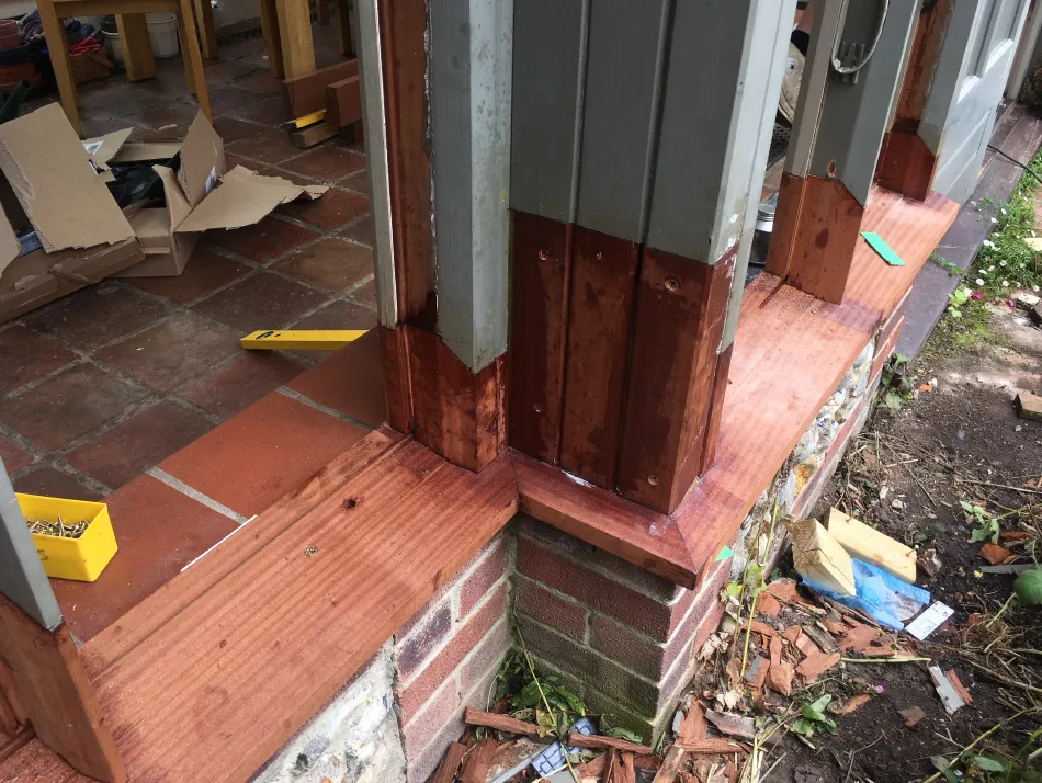 Close-up of wooden door frame and threshold with treated wood frame and brick base, surrounded by construction debris outdoors.