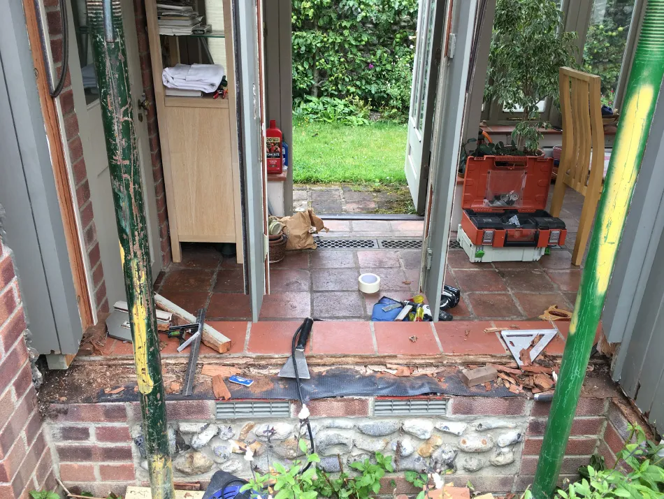 Open doorway under renovation with exposed bricks, tools, and supports, leading to a tiled floor and green garden outside.