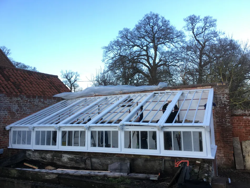 White wooden framed greenhouse structure under construction against a brick wall with leafless trees in the background.