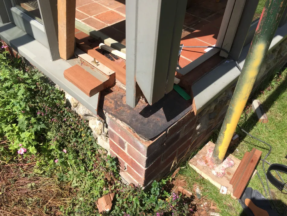 Close-up of construction detail showing a brick foundation with wooden framing and gray metal trim near a garden with green plants and flowers.
