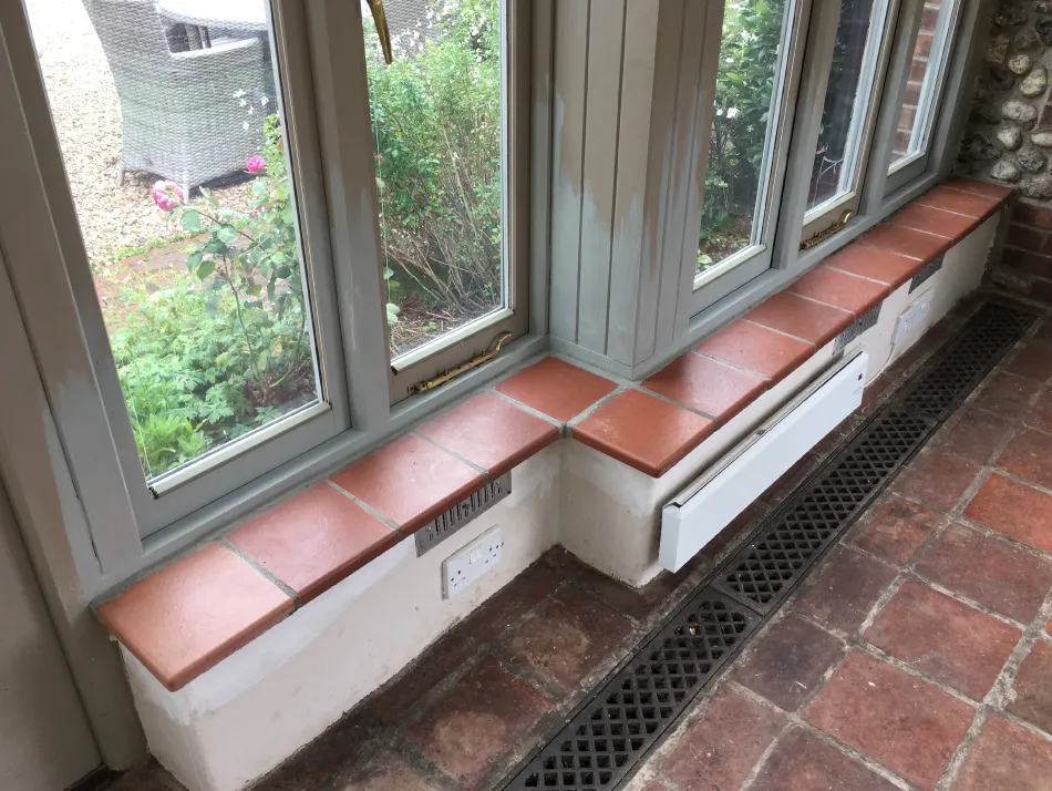Indoor window seating with terracotta tiles beneath large glass windows overlooking a garden, with a floor vent and wall heater below.