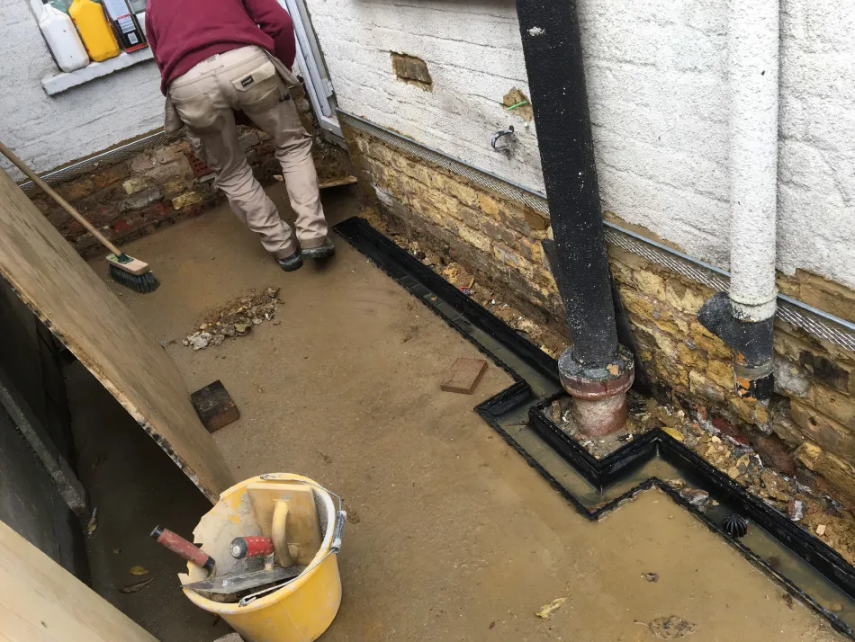 Worker in beige pants and maroon sweater installing drainage system along a building's brick foundation wall.