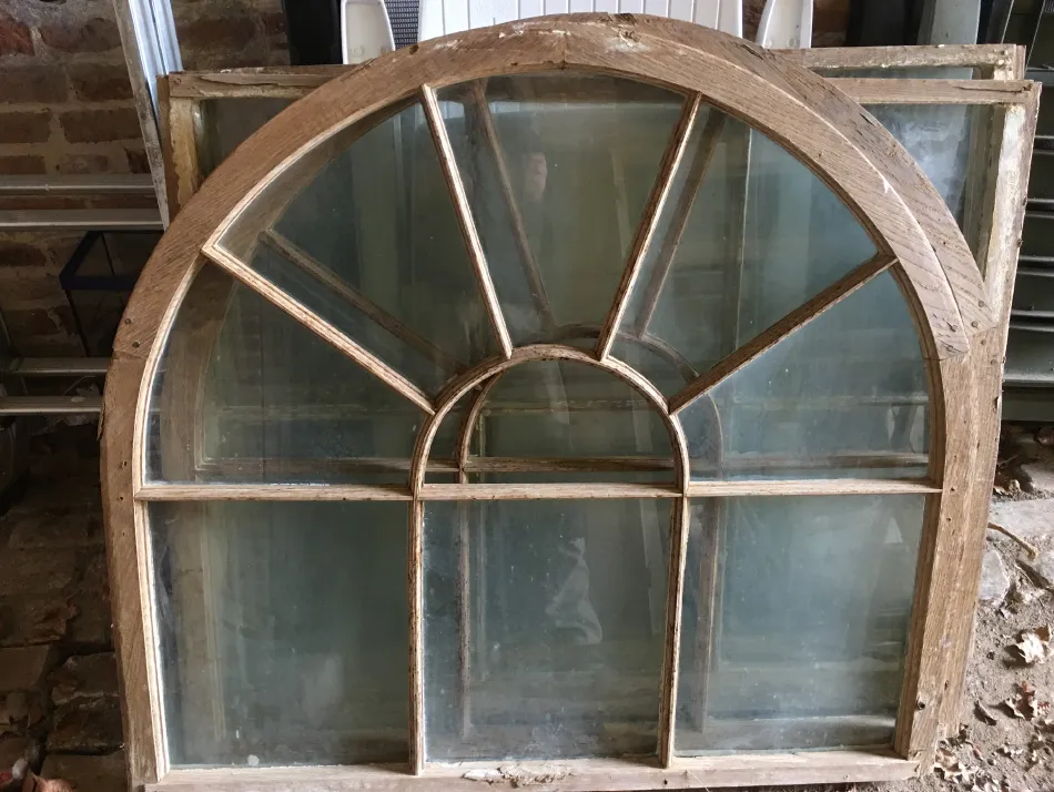 Arched vintage wooden window frame with multiple glass panes, resting against a wall in a rustic setting.