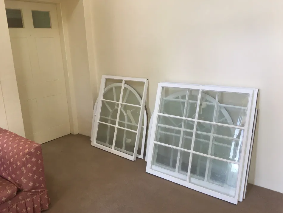 Two white-framed windows with multiple panes leaning against a beige wall on a carpeted floor near a closed door and a pink patterned sofa.