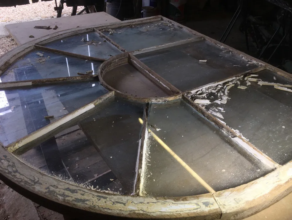 Round vintage window frame with multiple glass panes, some with damaged putty, resting on a surface in a work area.