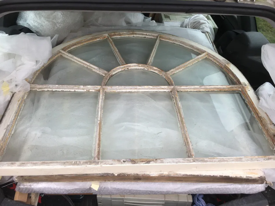 Arched window frame with peeling paint and six glass panes, lying flat inside a vehicle trunk surrounded by packing materials.