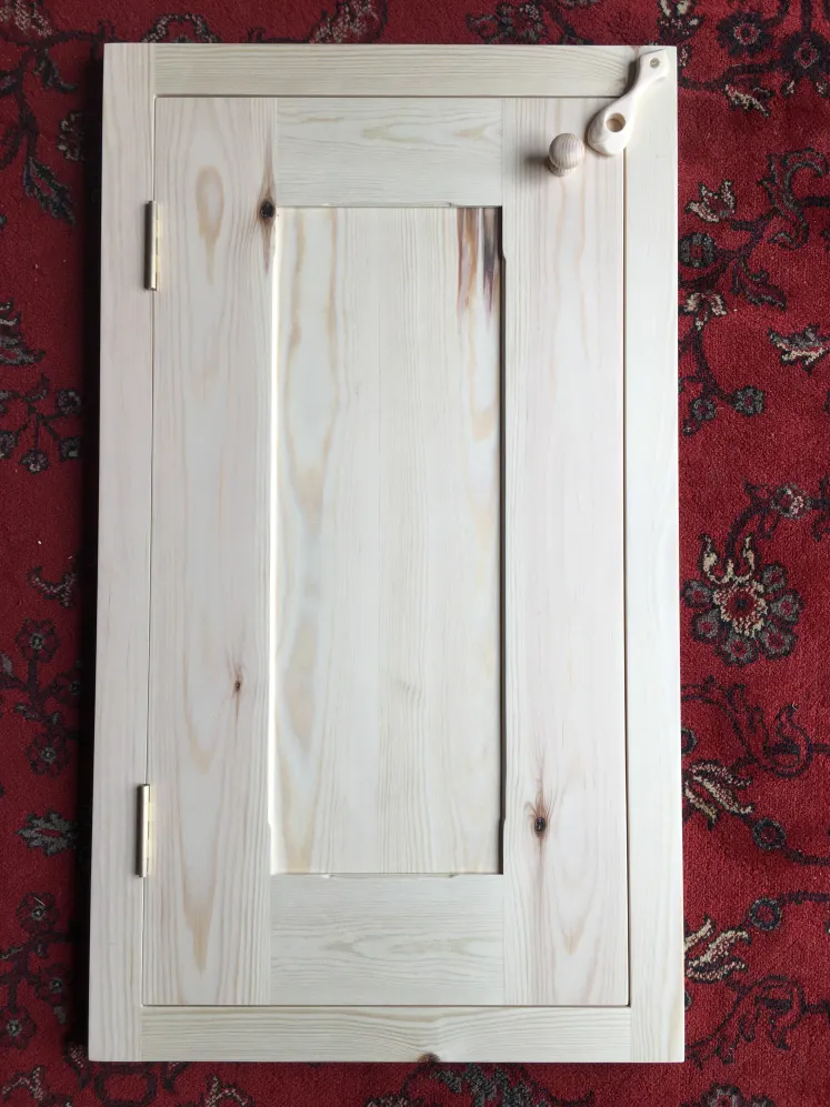 Unfinished wooden shaker-style cabinet door with brass hinges, a round wooden knob, and a wooden latch, placed on a red floral carpet.