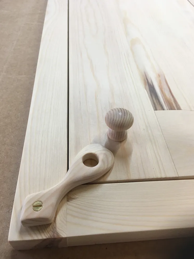 Unfinished light wood panel with a round wooden knob and a wooden pull handle with a hole and brass screw on its corner.