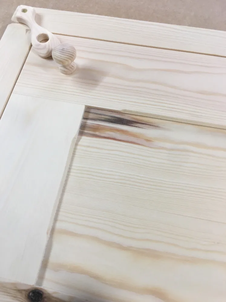 Close-up of an unfinished light wood Shaker cabinet door with a round wooden knob and a small wooden tool.