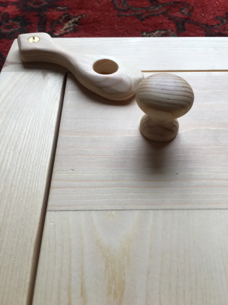 Unfinished light wood cabinet door with a round wooden knob and a separate wooden latch on a red patterned carpet background.