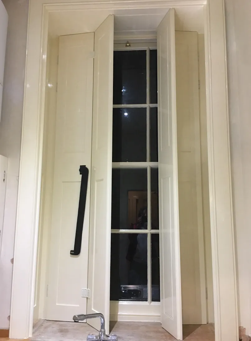 Cream-colored kitchen window with tall shutters, one slightly open, featuring a large black handle and a chrome faucet below.