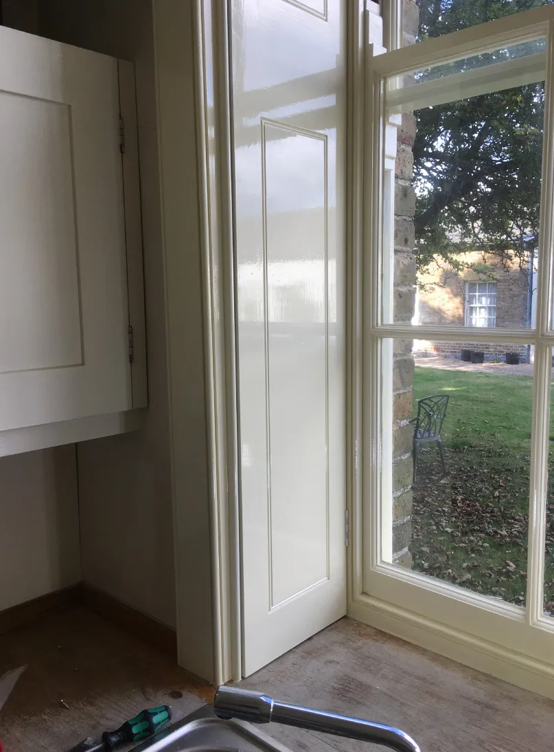 Partially open white wooden shutter beside glass window looking out onto a garden with lawn and metal chair.