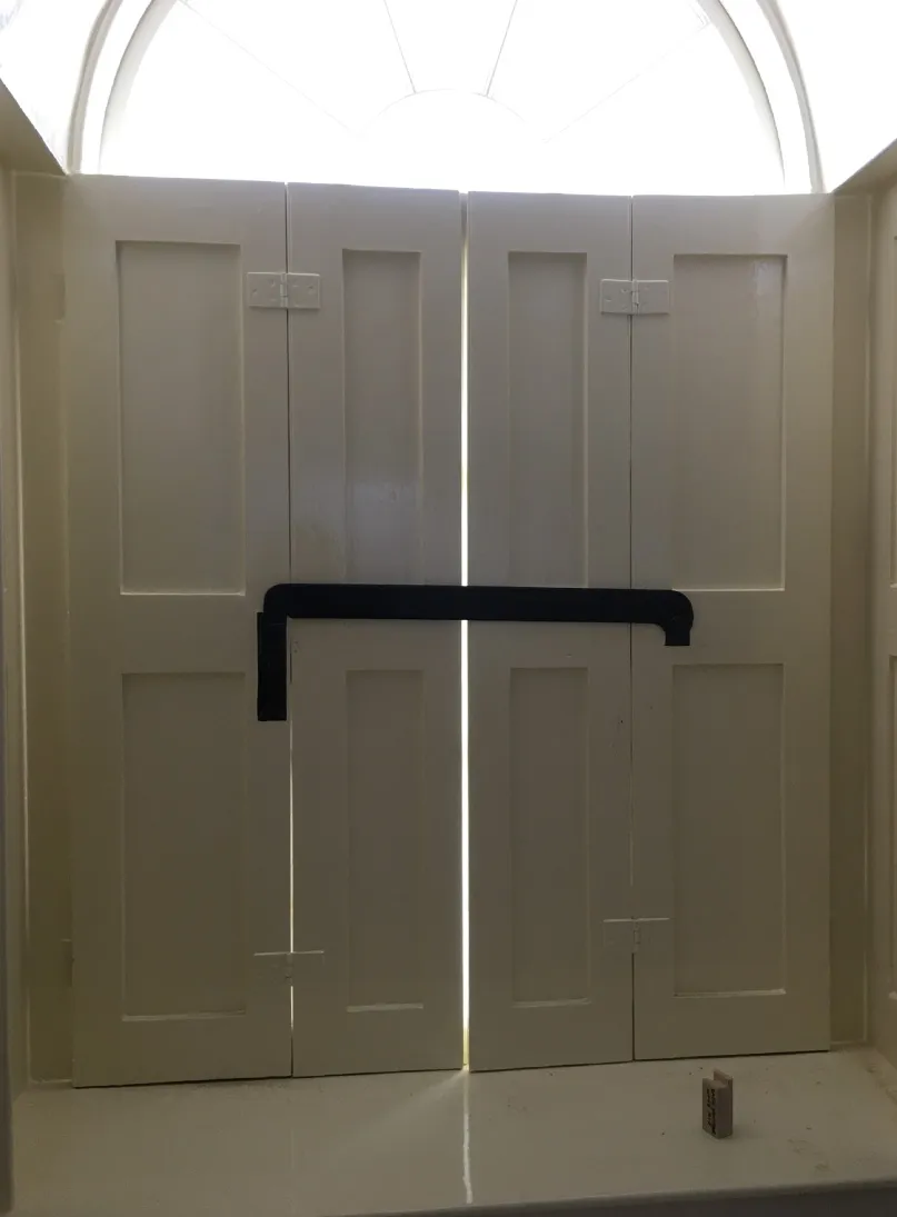 White wooden shutters with a central horizontal black bar lock, partially closed with light visible through the gaps and an arched window above.
