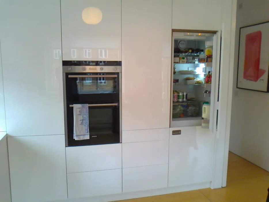 Modern white kitchen wall with built-in double oven and open refrigerator filled with food and drinks.