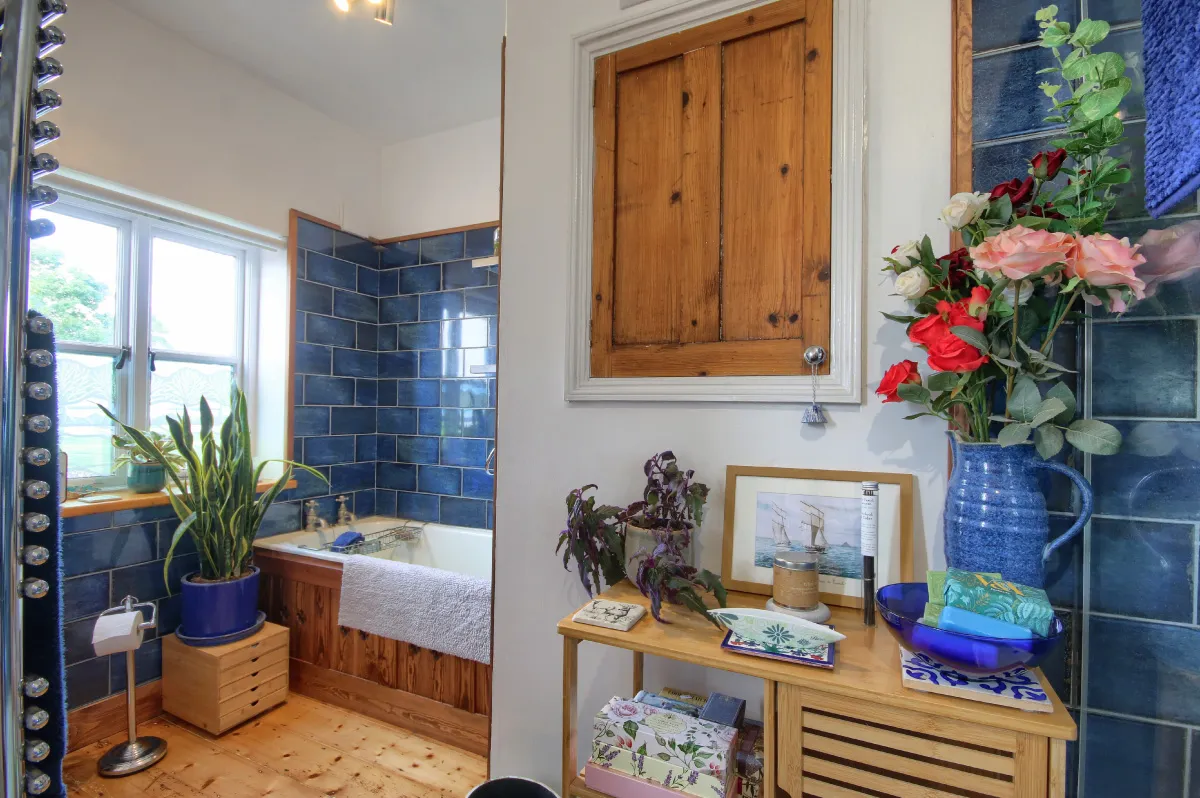 Bathroom with blue tiled walls, a wooden bathtub, potted plants by the window and on a wooden cabinet topped with flowers and decorative items.