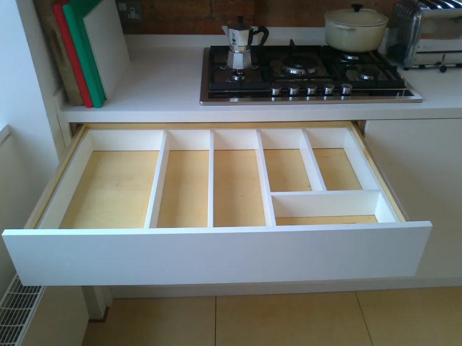 Open kitchen drawer with white wooden organizers and compartments under a countertop with a gas stove, moka pot, and cookware.