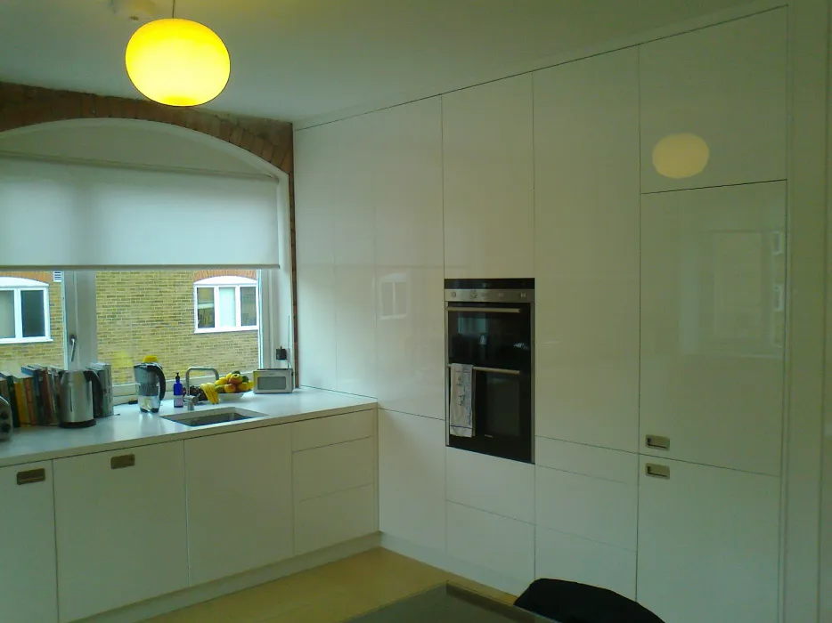 Modern kitchen with white glossy cabinets, built-in oven, sink under a window with a white roller blind, and a round yellow ceiling light.