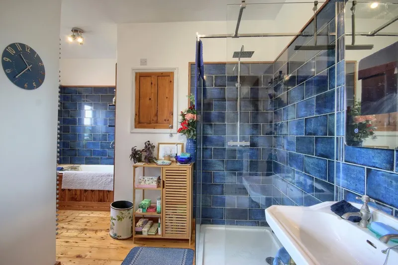 Bright bathroom with blue tiled walk-in shower, white sink, wooden shelf unit with toiletries, and wooden floor.