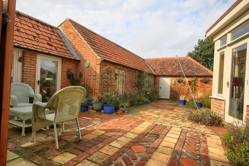 Cozy brick courtyard with terracotta tiled roof, wicker chairs, table, and various potted plants.