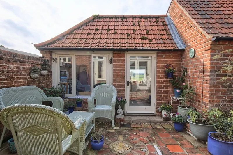 Cozy brick patio with red tile roof, white wicker chairs and table, and various potted plants around.