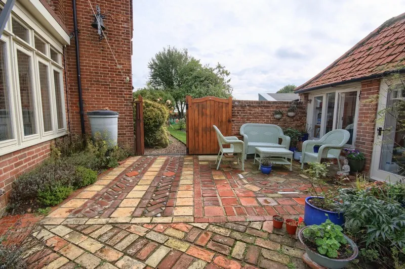 Patio area with patterned brick flooring, outdoor seating set, potted plants, and a wooden gate opening to a garden.