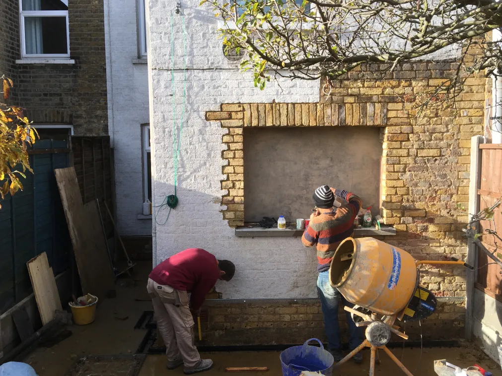 Two workers renovating a brick wall and window recess, with one measuring and the other applying materials near a yellow cement mixer.