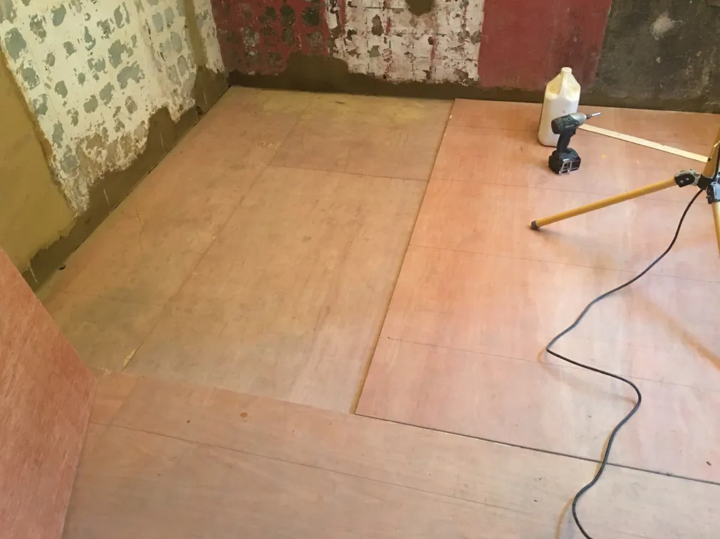 Room under refurbishment with plywood sheets partially installed on the floor and construction tools including a drill, a plastic bottle, and a tripod light stand.