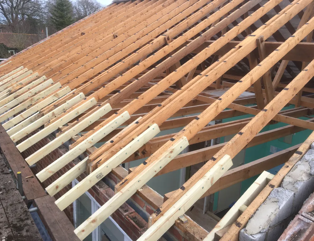 Roof structure with wooden rafters and new light-colored timber pieces being installed on an angled roof frame.