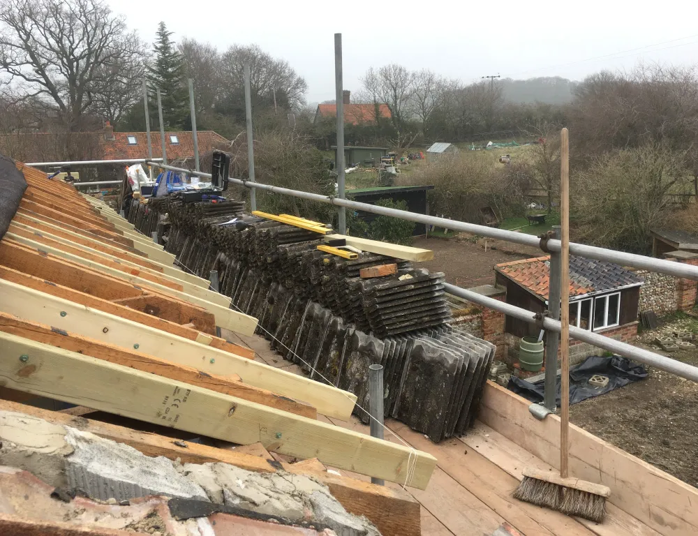 Roof under construction with wooden beams and stacked tiles, scaffolding, broom, and a rural landscape in the background.