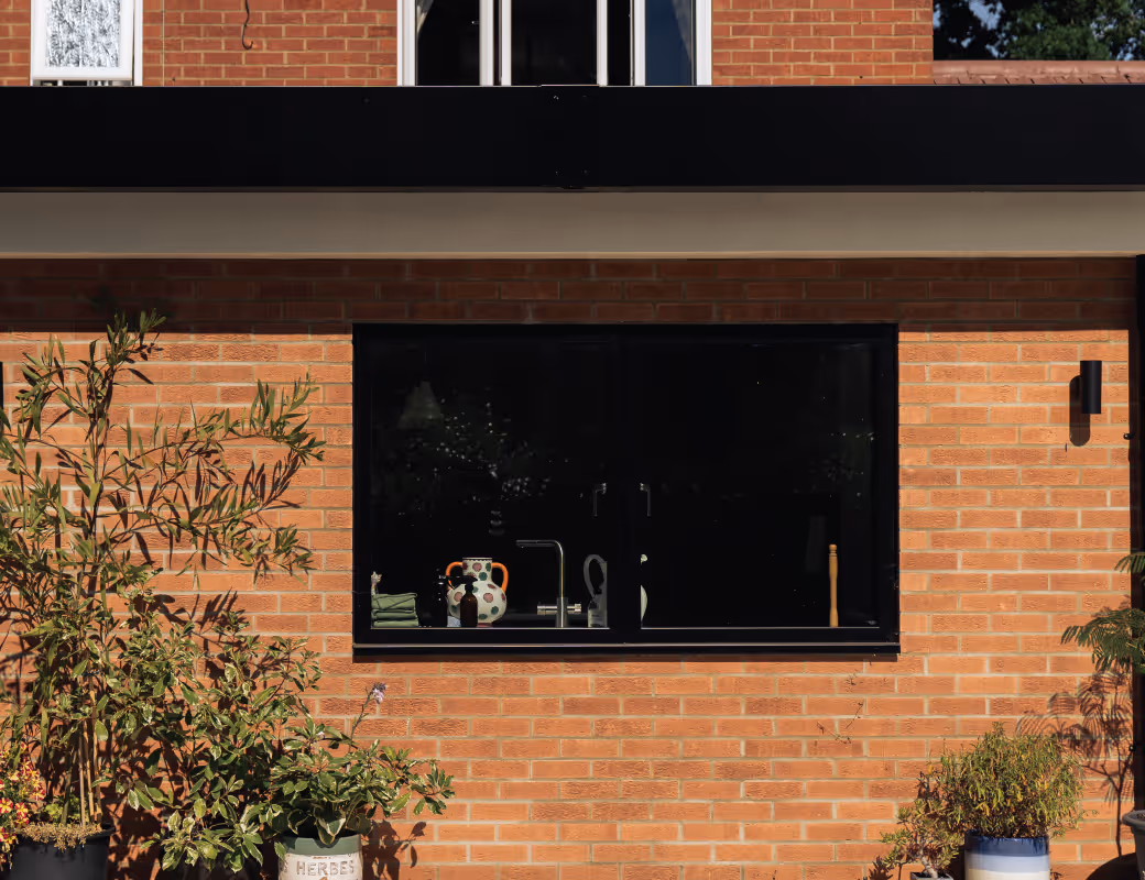 Worker in beige pants and maroon sweater installing drainage system along a building's brick foundation wall.