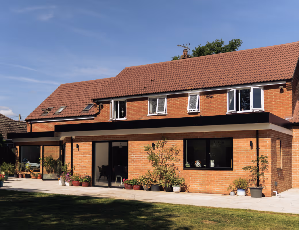 Worker in beige pants and maroon sweater installing drainage system along a building's brick foundation wall.