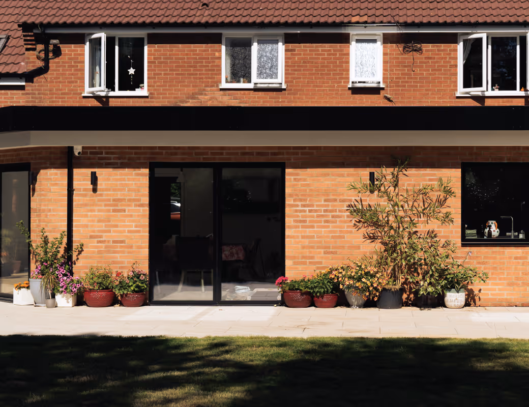 Worker in beige pants and maroon sweater installing drainage system along a building's brick foundation wall.