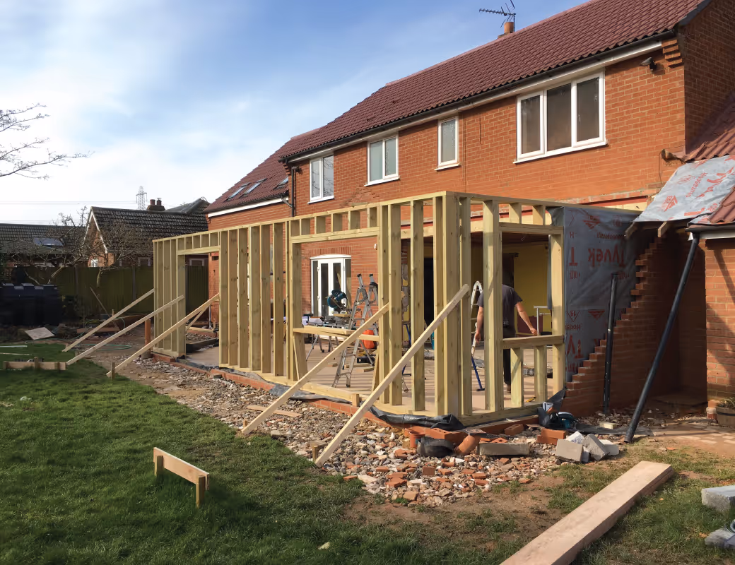 Worker in beige pants and maroon sweater installing drainage system along a building's brick foundation wall.