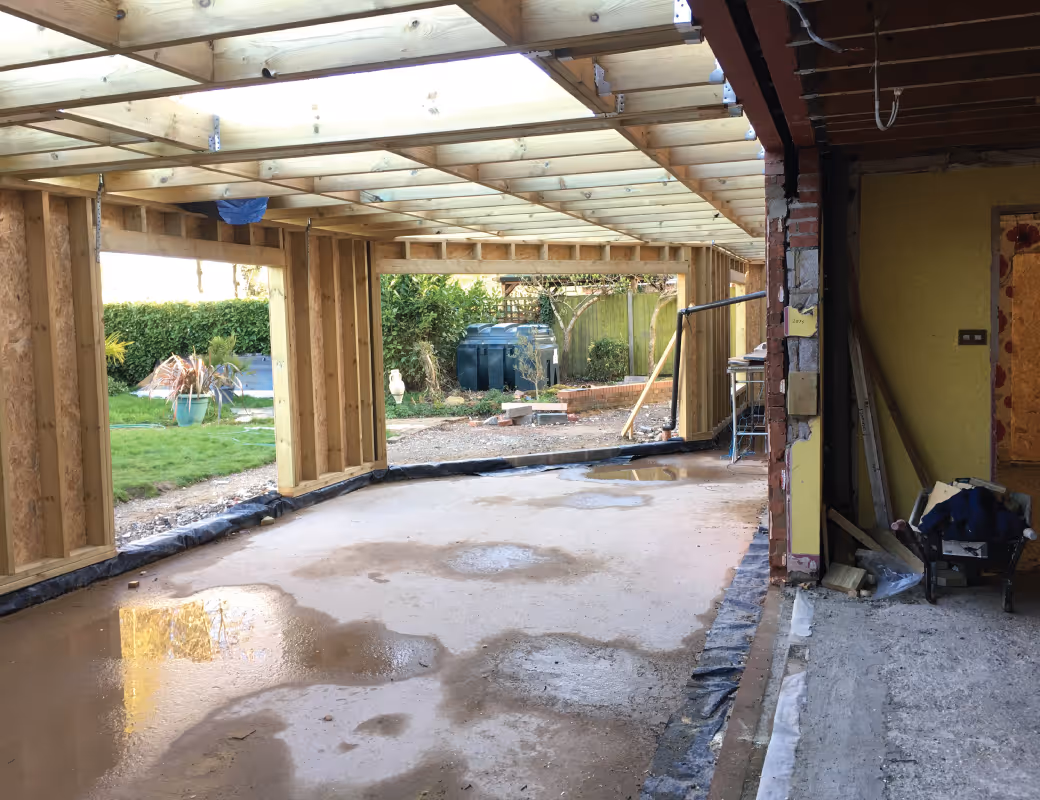 Worker in beige pants and maroon sweater installing drainage system along a building's brick foundation wall.