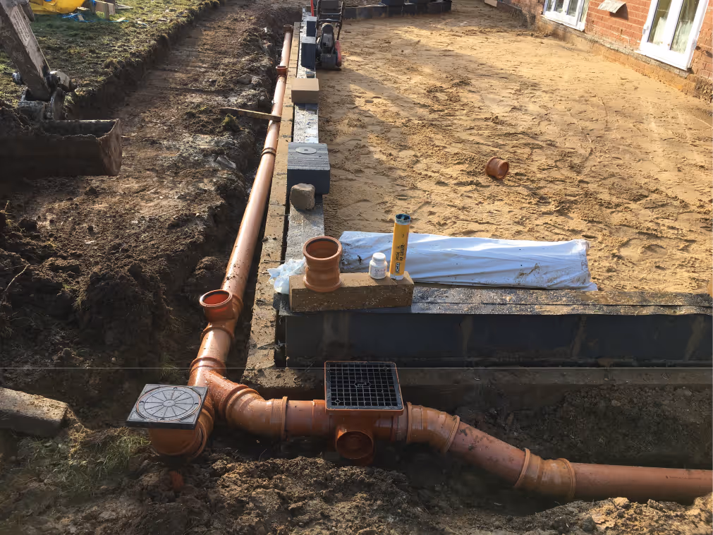 Worker in beige pants and maroon sweater installing drainage system along a building's brick foundation wall.