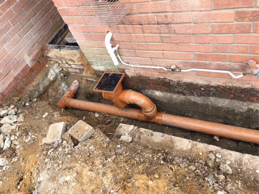 Worker in beige pants and maroon sweater installing drainage system along a building's brick foundation wall.