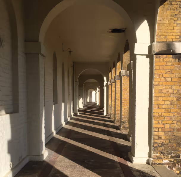 Sunlit arched corridor with white columns on the left and brick walls on the right casting long shadows on the floor.