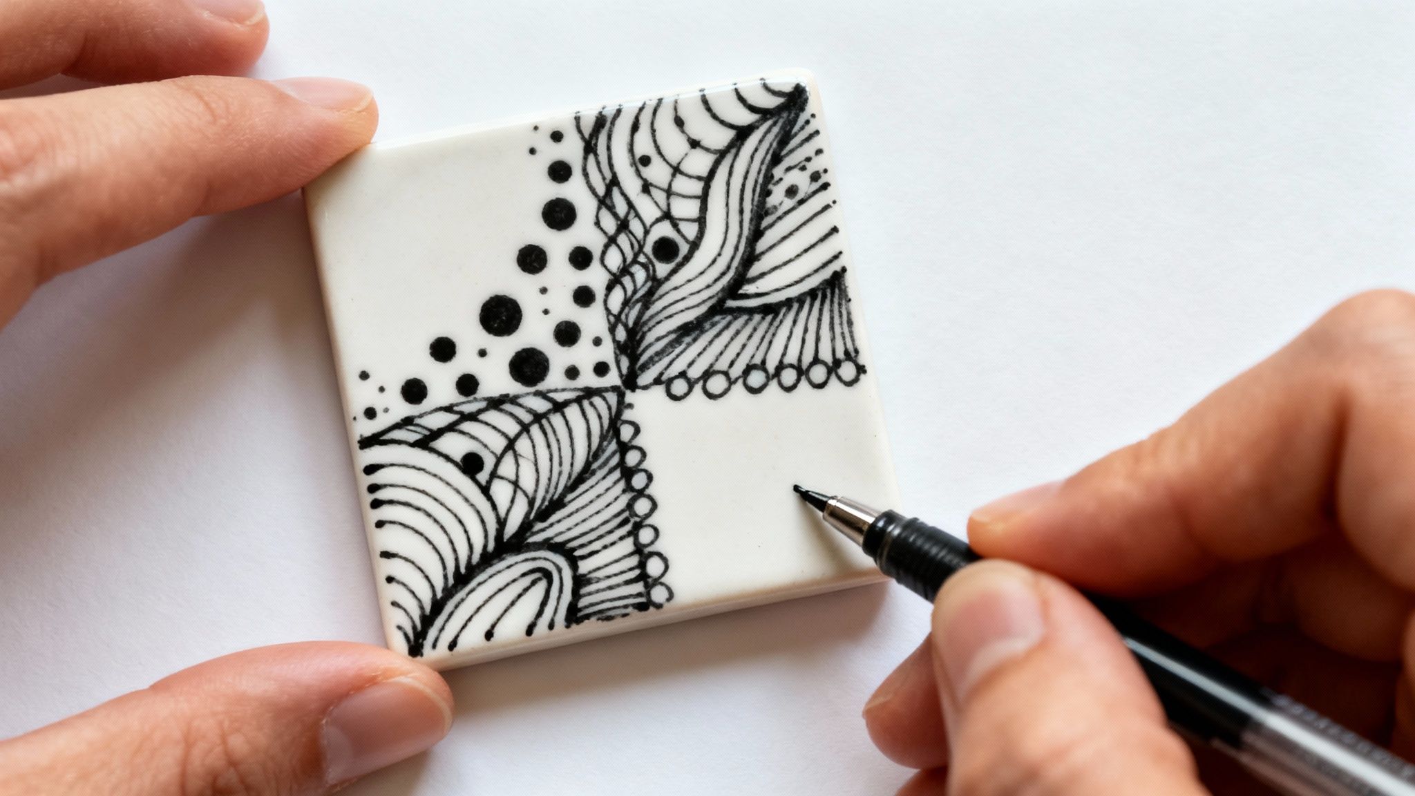 Close-up of hands drawing intricate black and white Zentangle patterns on a small white tile.