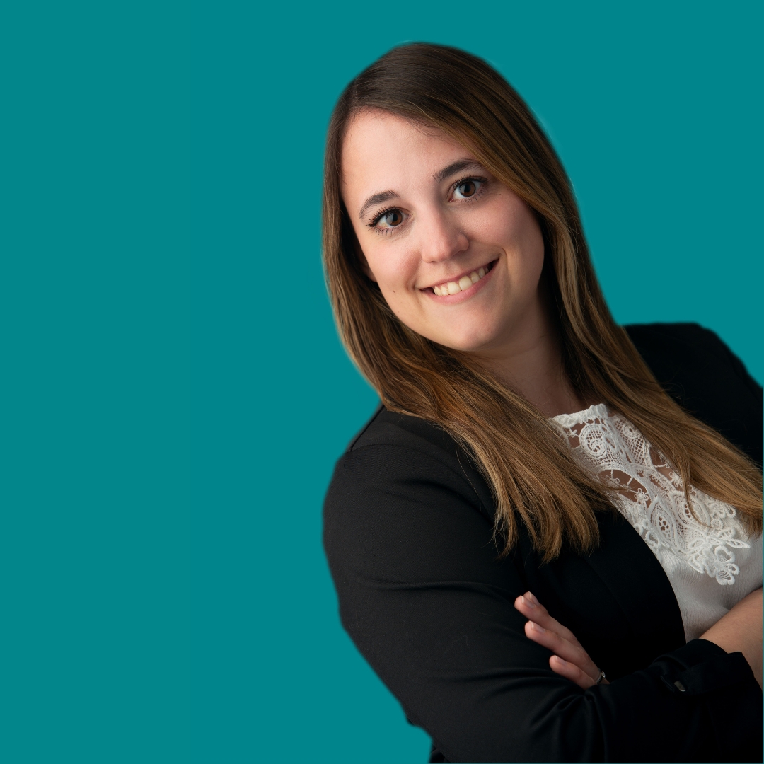 Porträt einer brünetten, freundlichen Frau, die mit verschränkten Armen lächelt. Der Hintergrund ist türkis.