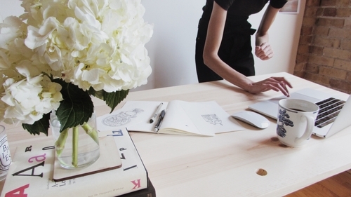 Person working on a laptop at a wooden desk with a white floral bouquet, sketches, pens, and a coffee mug.