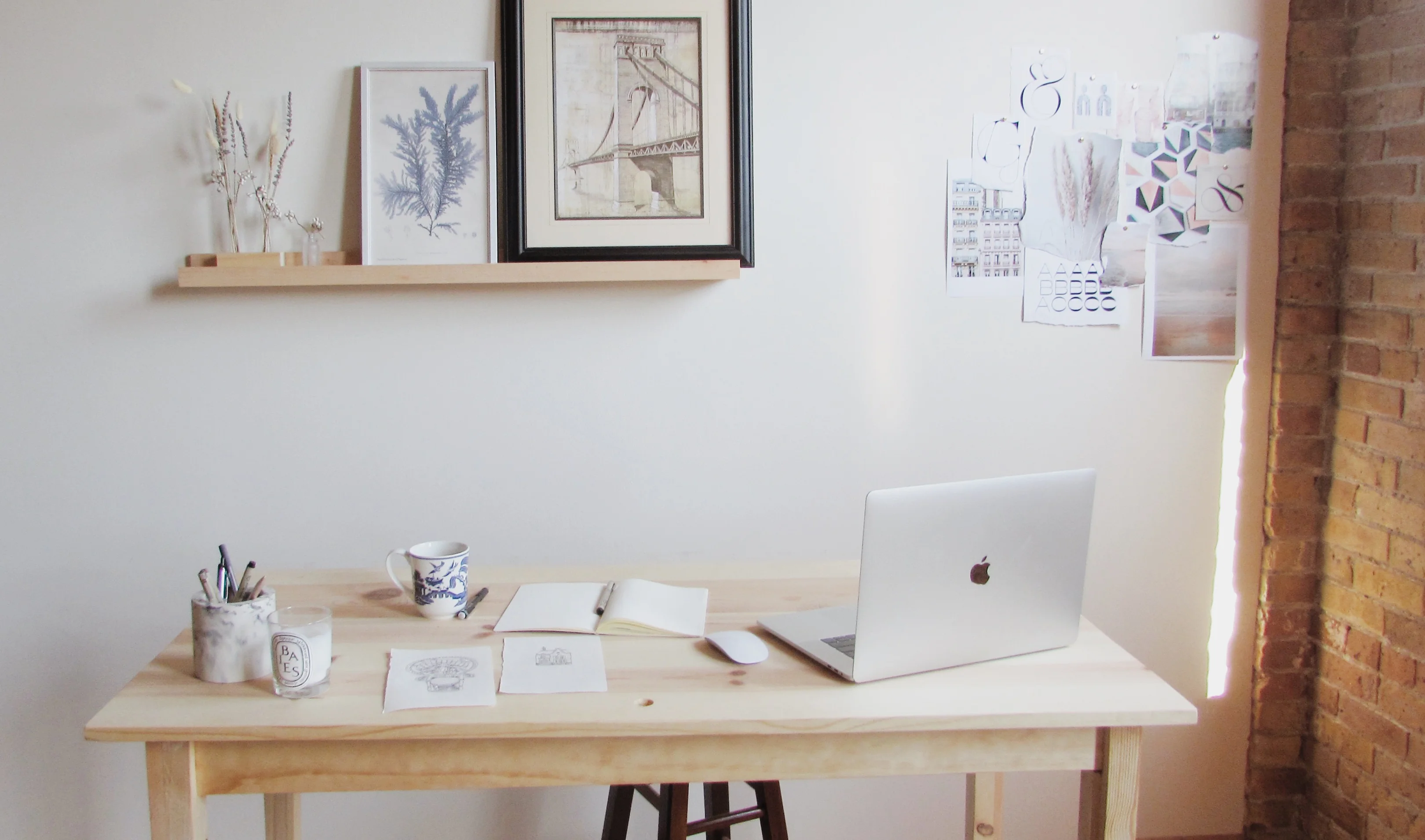 Minimal wooden desk with a laptop, mouse, notebook, pen, cup, drawings, and pencil holder under a shelf with framed artwork and dried plants.