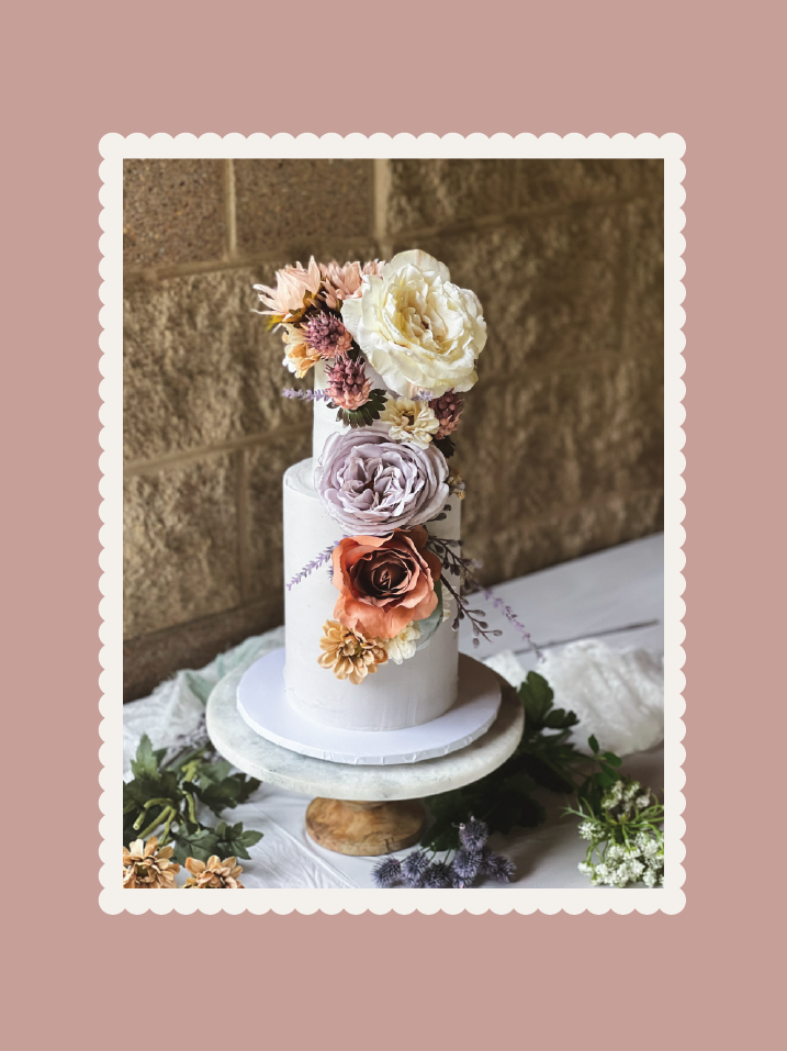 Two-tier white cake adorned with large pastel-colored flowers on a marble cake stand with greenery around it.