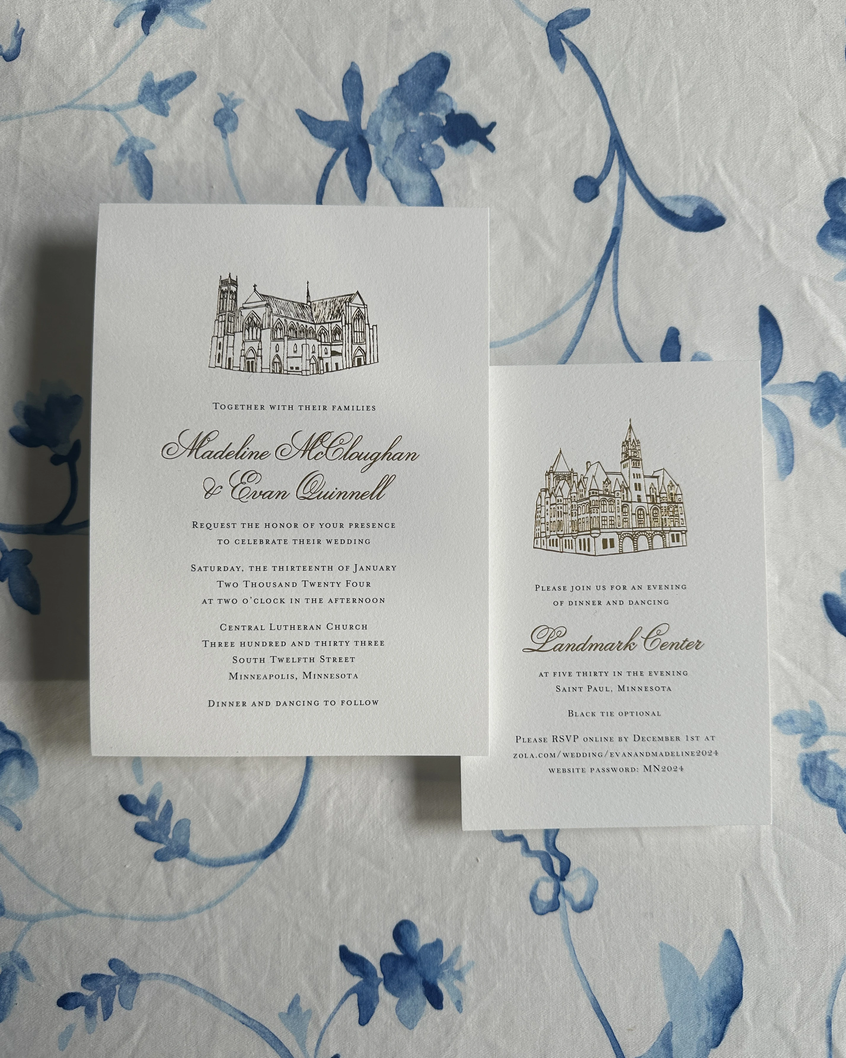 Two white wedding invitations with gold building illustrations on blue floral fabric.