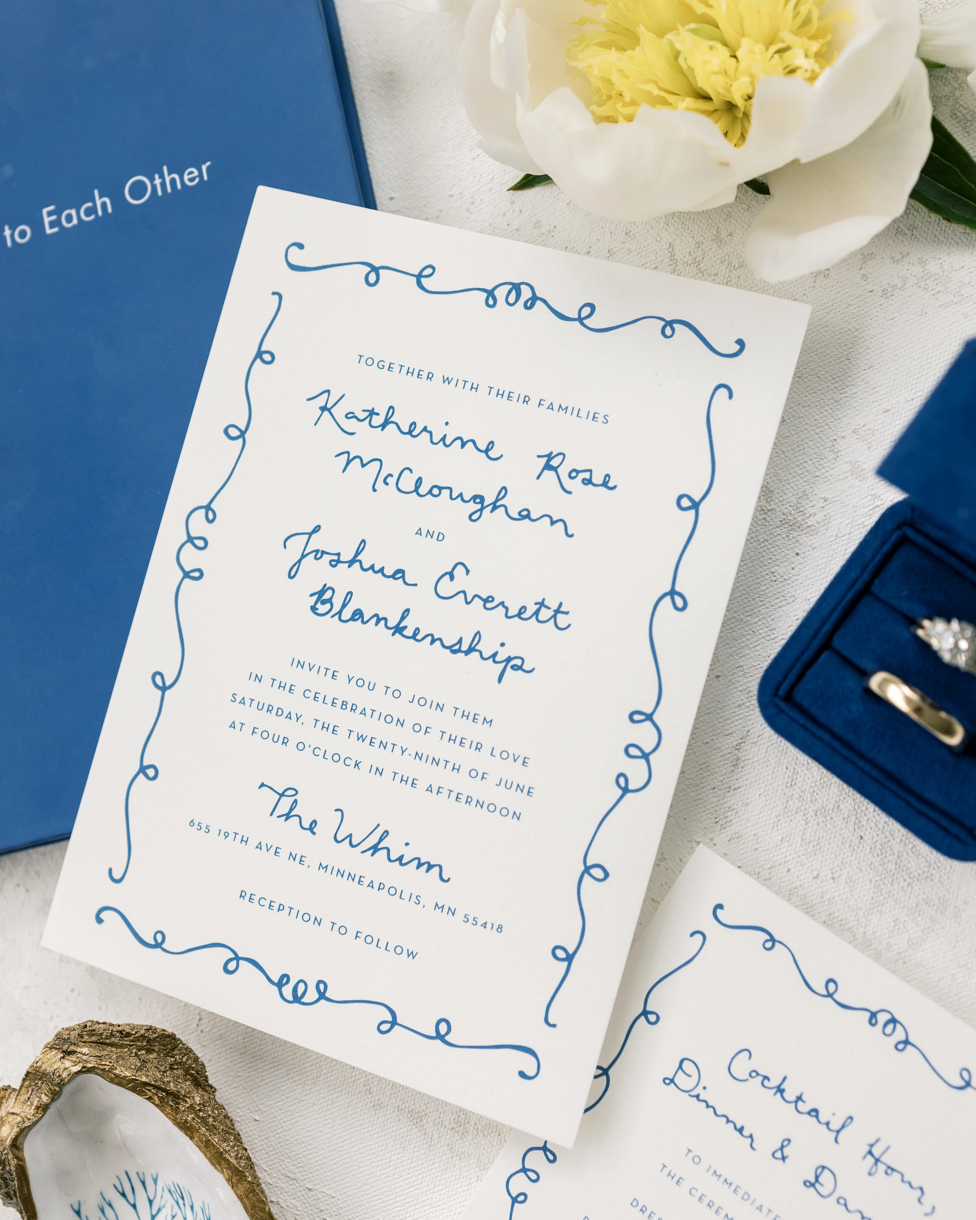 Wedding invitation suite with navy blue book, rings in blue box, white flower, and decorative shell.