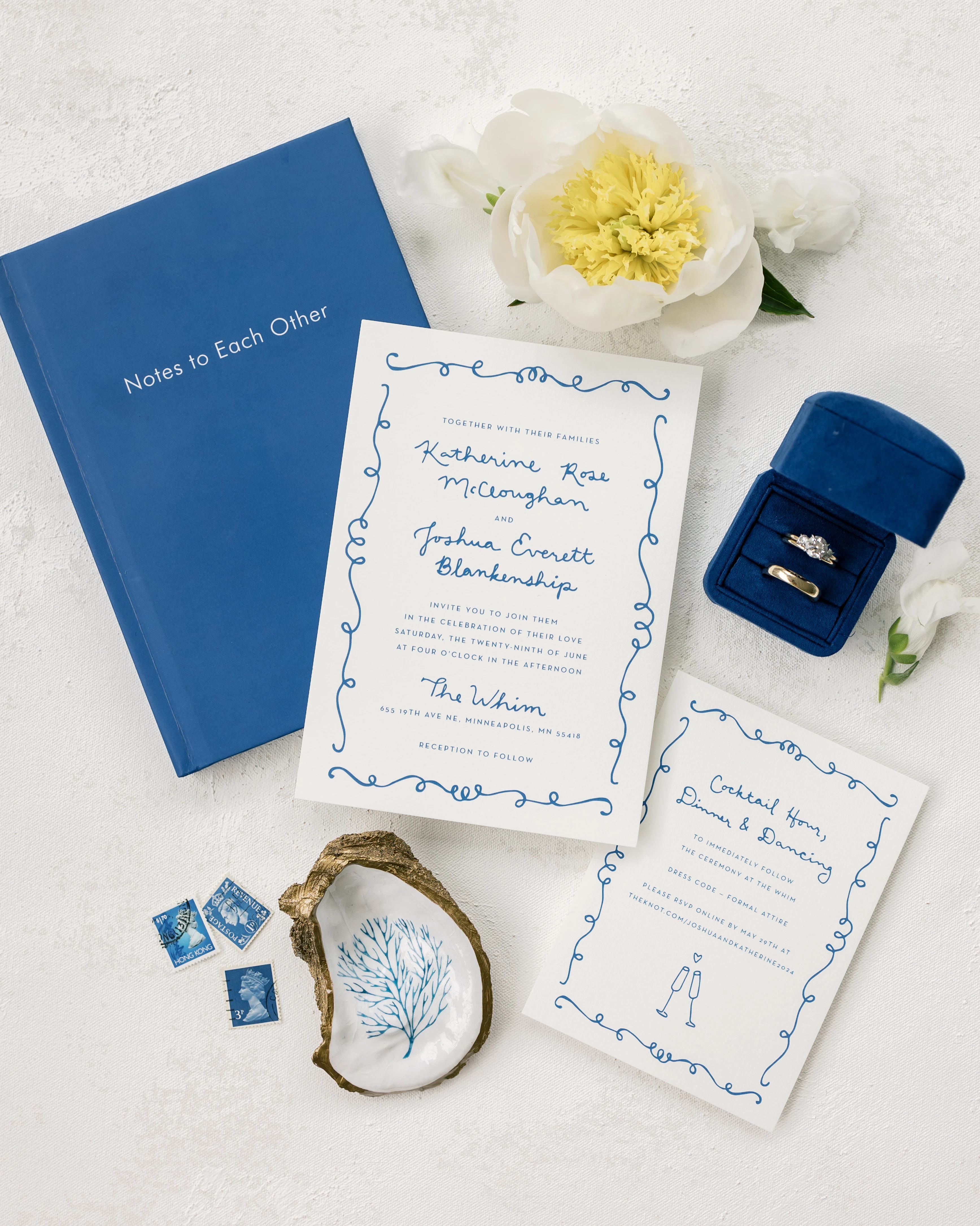 Wedding invitation with blue cursive text, surrounded by a blue squiggly border, beside a flower and ring box.