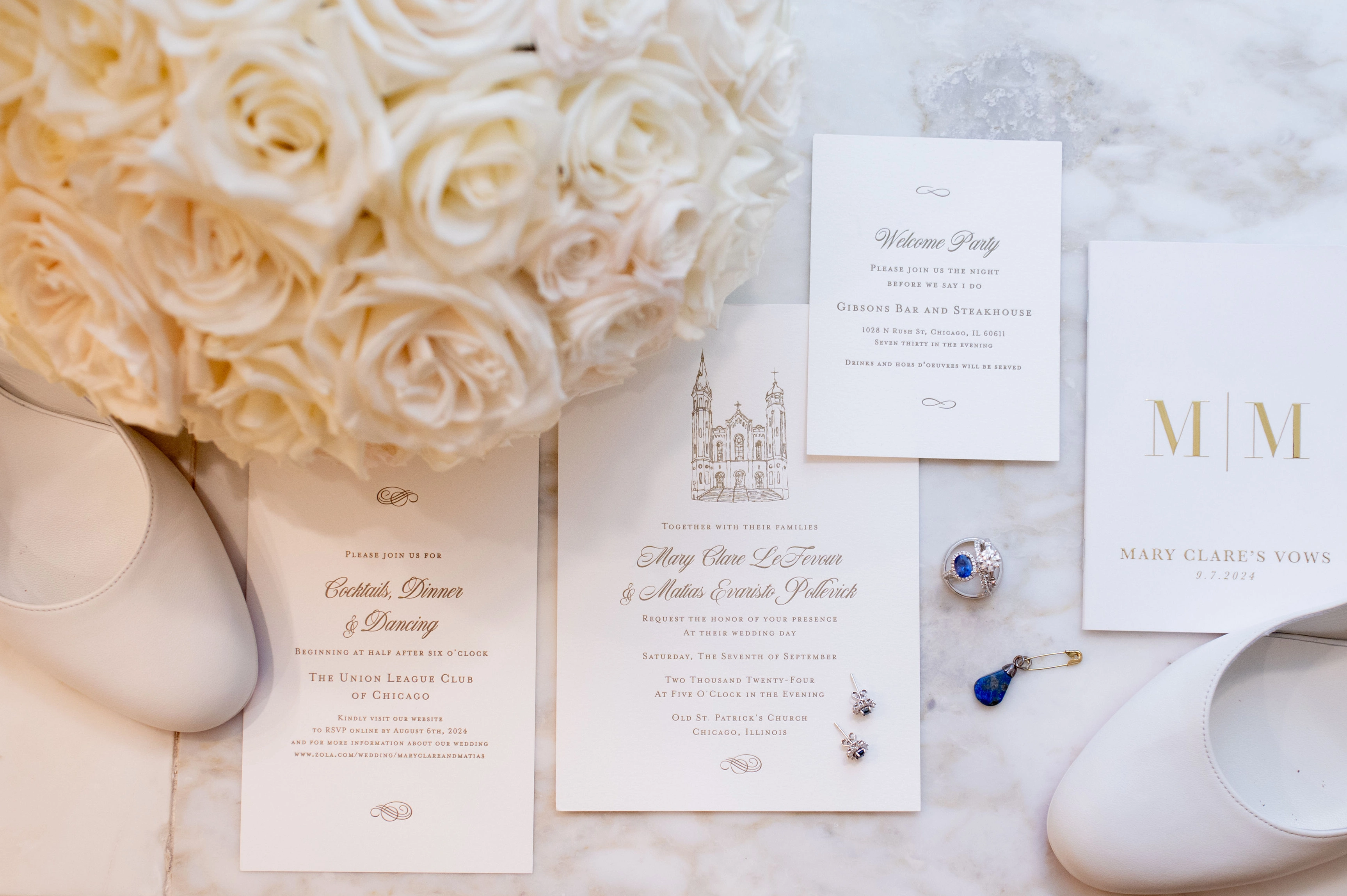 Blue floral wedding shoes, pearl clutch, rings, flower, and wedding invitations on a light blue cloth.