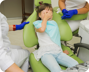 Young girl sitting nervously in a dental chair covering her eyes with one hand while a dentist and assistant wearing gloves talk to her.