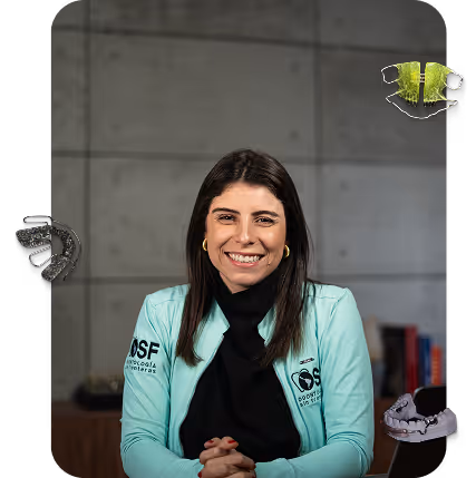 Smiling woman with dark hair wearing a turquoise jacket with logo, sitting with clasped hands against a gray tiled wall background.