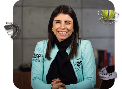 Smiling woman in a light blue lab coat with dental company logos, sitting with hands clasped in front of a gray wall background.